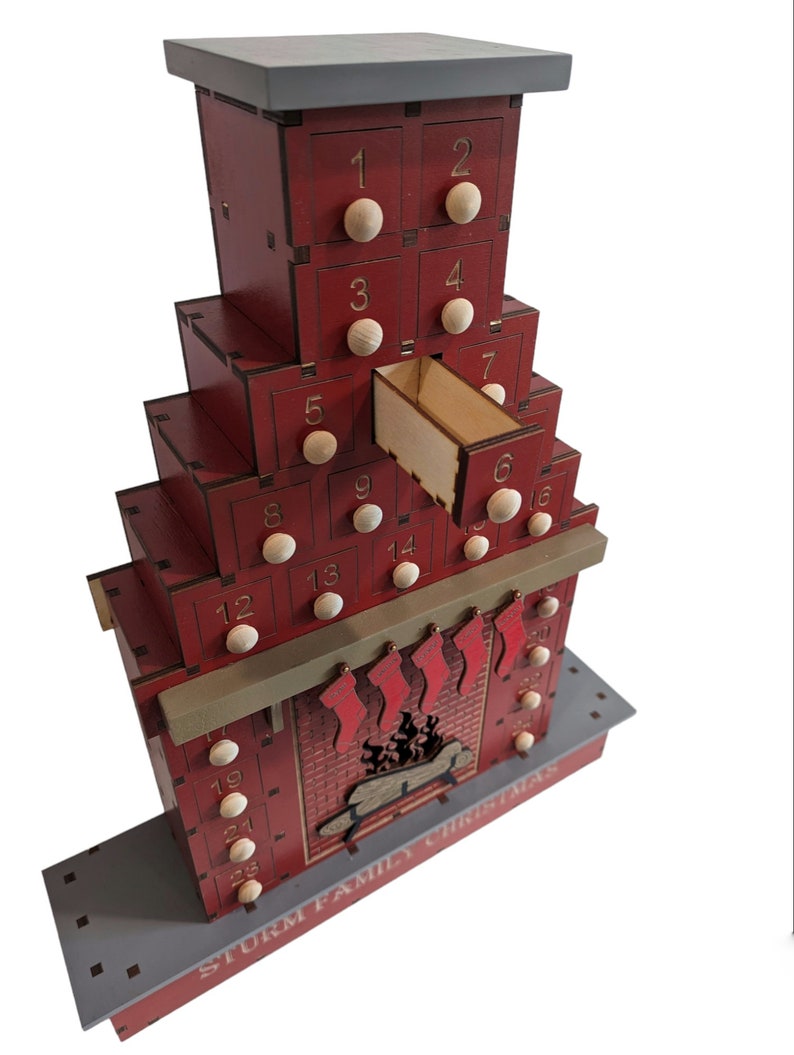 May include: A red wooden Christmas advent calendar shaped like a chimney with 25 drawers. The drawers are numbered 1 through 25. The calendar has a gray roof and a gray base. The text "STURM FAMILY CHRISTMAS" is printed on the base.