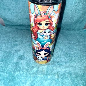May include: A stainless steel tumbler with a clear lid, featuring colourful illustrations of cartoon characters with bunny ears. The design includes a red-haired character holding a blue bunny and a dark-haired character with a white bunny.