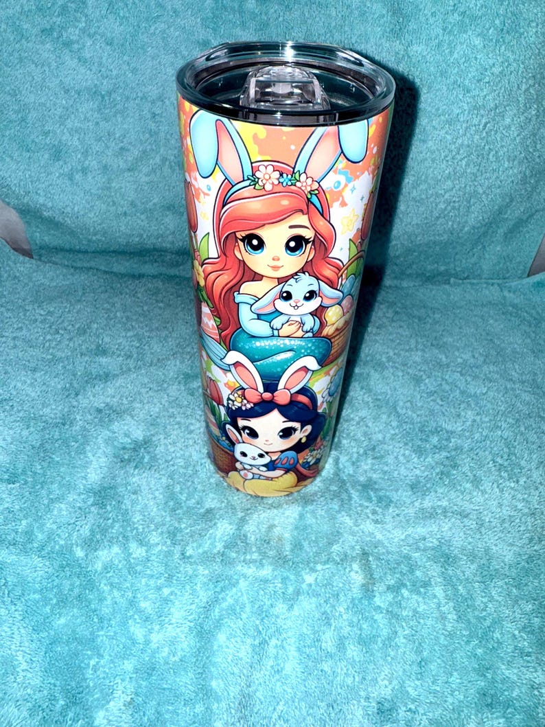 May include: A stainless steel tumbler with a clear lid, featuring colourful illustrations of cartoon characters with bunny ears. The design includes a red-haired character holding a blue bunny and a dark-haired character with a white bunny.