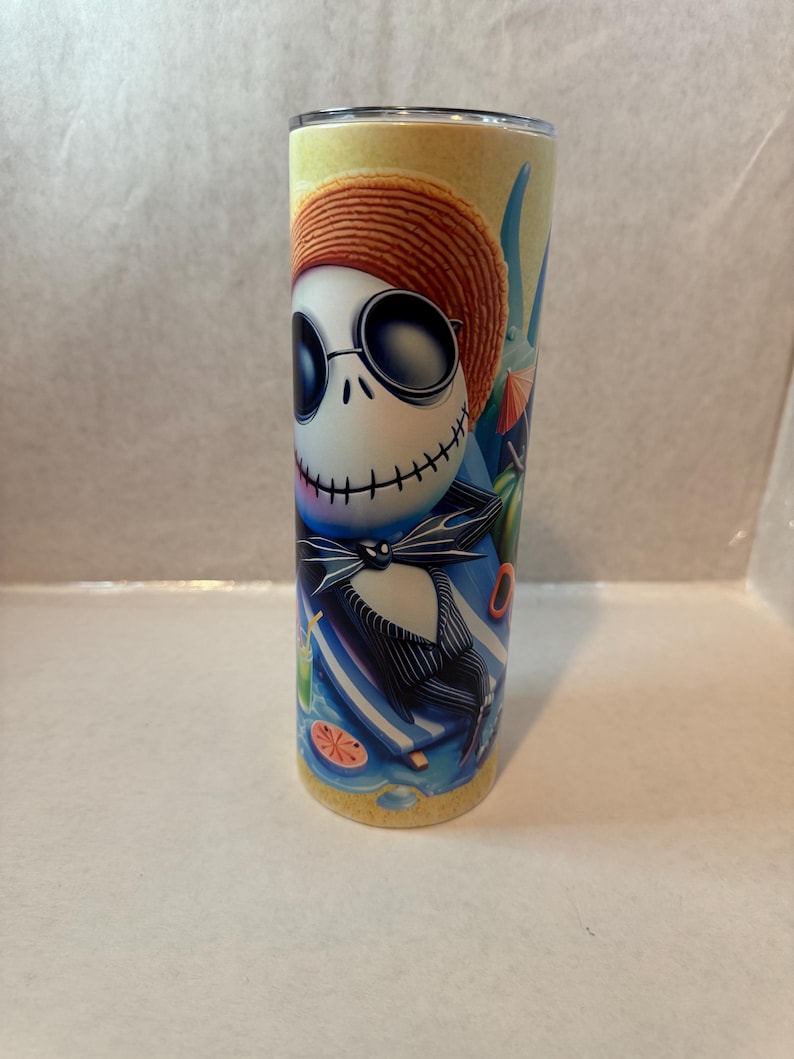 K&ouml;nnte beinhalten: Ein Edelstahlbecher mit einer farbenfrohen Illustration von Jack Skellington aus The Nightmare Before Christmas. Jack ist mit Sonnenbrille und Strohhut dargestellt, wie er sich an einem Strand mit einem Cocktail entspannt. Der Becher ist zylindrisch und hat einen silbernen Deckel.