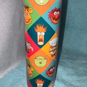 May include: A stainless steel tumbler featuring a colorful geometric pattern with various Muppet characters. The design includes faces of Fozzie Bear, Animal, Beaker, and others. The tumbler has a silver lid and is designed to keep beverages cold or hot.