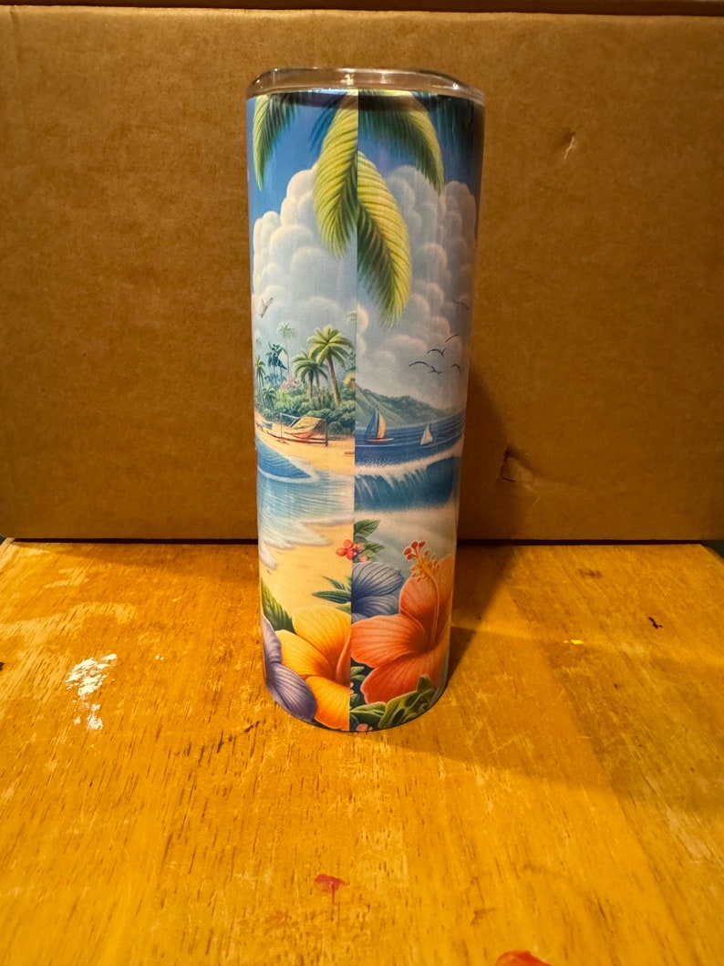 May include: A stainless steel tumbler with a tropical beach scene design. The design features palm trees, a beach, ocean waves, and colorful hibiscus flowers.