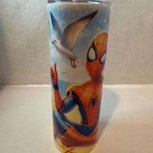 May include: A stainless steel tumbler with a colorful design featuring Spiderman sitting on a beach with a seagull flying overhead. The design includes a starfish and palm trees.