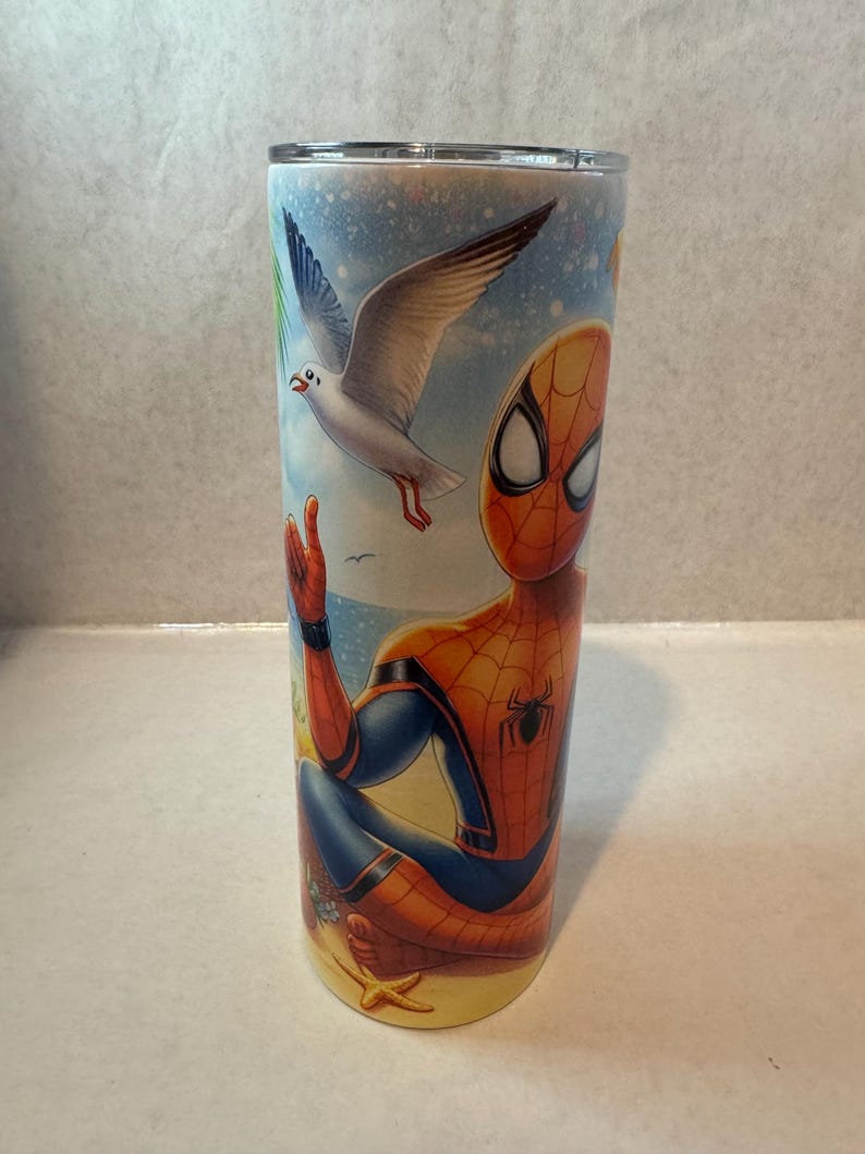 May include: A stainless steel tumbler with a colorful design featuring Spiderman sitting on a beach with a seagull flying overhead. The design includes a starfish and palm trees.