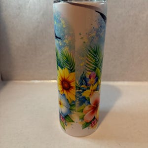 May include: A white stainless steel tumbler with a tropical floral design. The design features yellow sunflowers, pink hibiscus flowers, blue flowers, and green palm leaves on a white background.