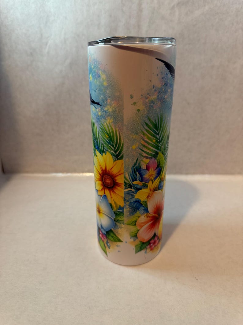 May include: A white stainless steel tumbler with a tropical floral design. The design features yellow sunflowers, pink hibiscus flowers, blue flowers, and green palm leaves on a white background.