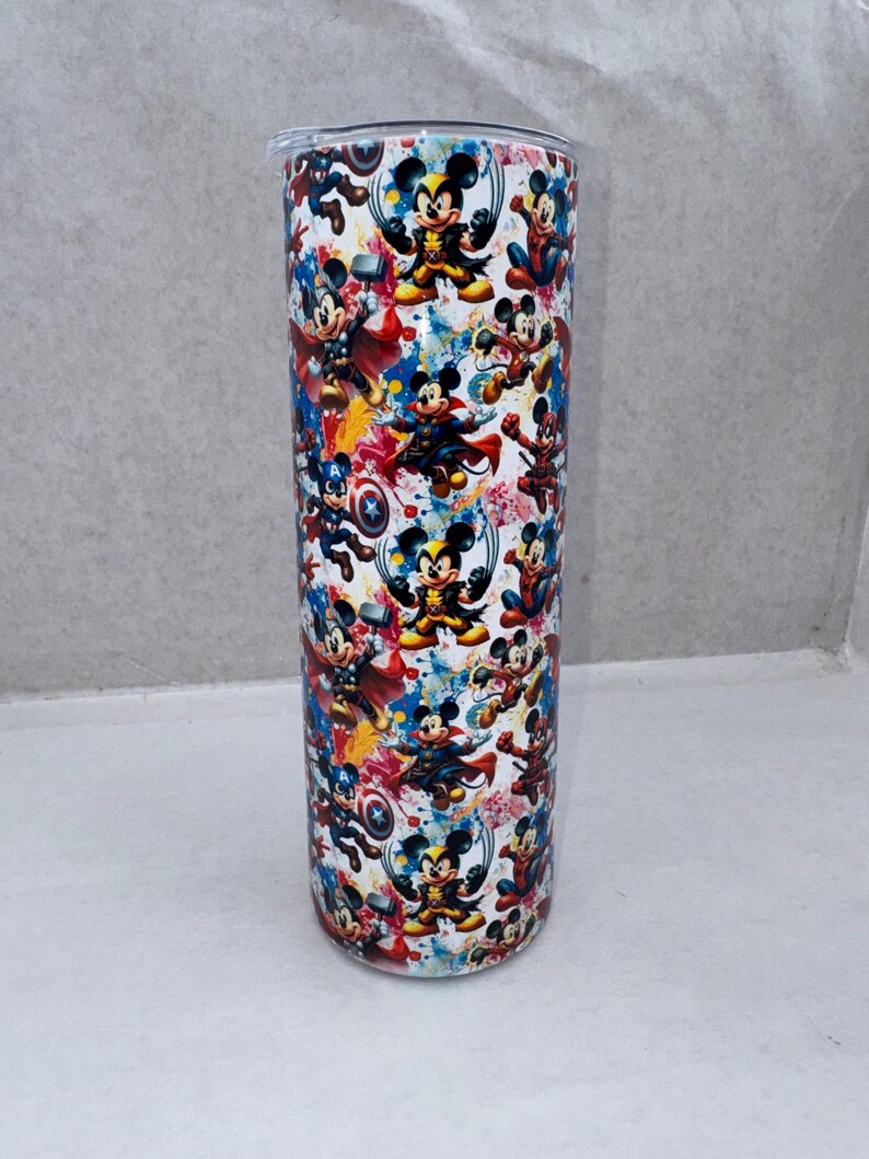 May include: A cylindrical tumbler featuring a repeating pattern of Mickey Mouse dressed as various superheroes, including Wolverine, Thor, Captain America, and Spider-Man, set against a white background with colorful paint splatter accents.