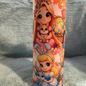 May include: A tall, cylindrical tumbler with cartoon illustrations of two princesses wearing bunny ears, holding bunnies, and surrounded by Easter eggs and baskets. The design has a pastel colour scheme with orange, pink, and blue hues.
