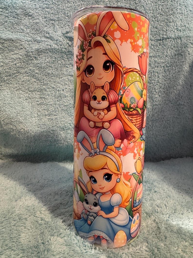 May include: A tall, cylindrical tumbler with cartoon illustrations of two princesses wearing bunny ears, holding bunnies, and surrounded by Easter eggs and baskets. The design has a pastel colour scheme with orange, pink, and blue hues.