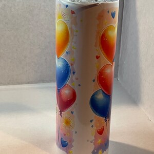 Pode incluir: Um copo de a&ccedil;o inoxid&aacute;vel branco com um design colorido de bal&otilde;es em forma de cora&ccedil;&atilde;o. Os bal&otilde;es s&atilde;o vermelhos, azuis, amarelos e laranja. O fundo &eacute; rosa claro com estrelas brancas e um efeito aquarela.