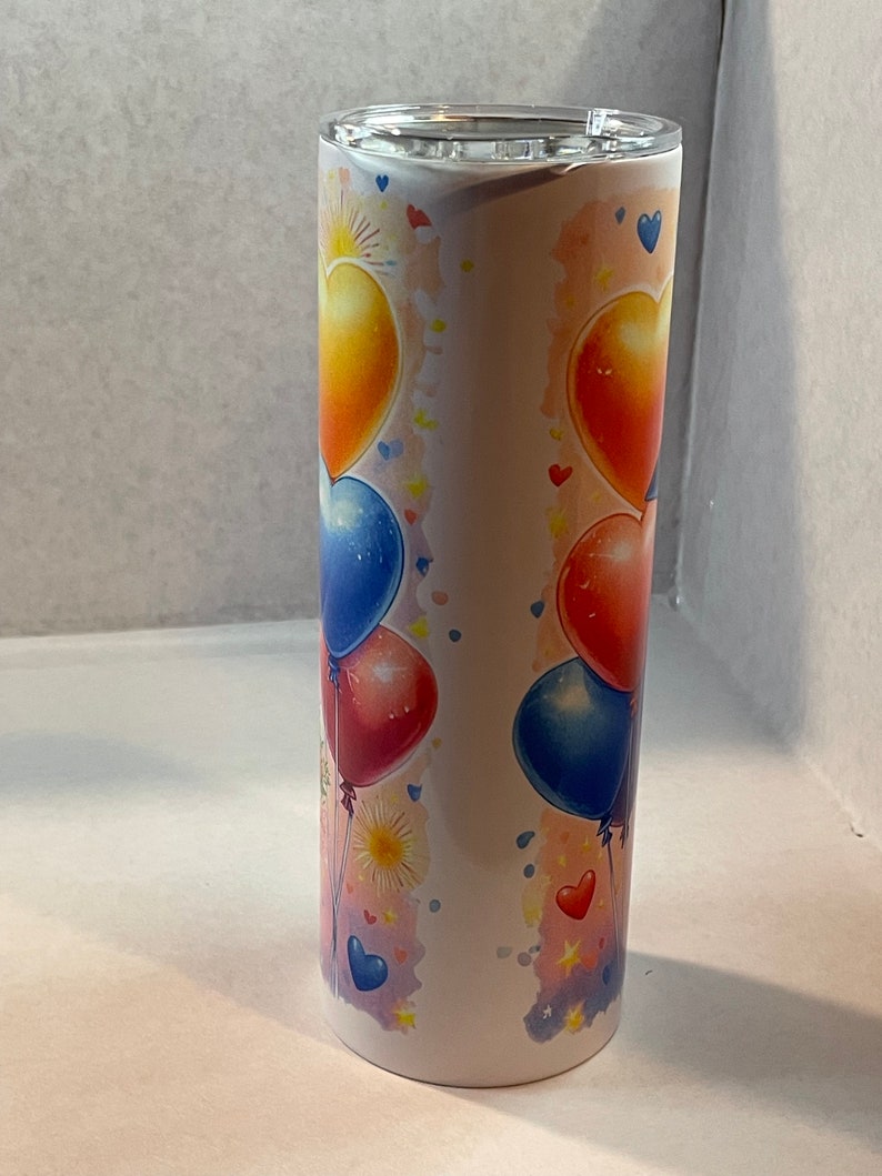 Pode incluir: Um copo de a&ccedil;o inoxid&aacute;vel branco com um design colorido de bal&otilde;es em forma de cora&ccedil;&atilde;o. Os bal&otilde;es s&atilde;o vermelhos, azuis, amarelos e laranja. O fundo &eacute; rosa claro com estrelas brancas e um efeito aquarela.