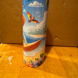 May include: A stainless steel tumbler with a colorful tropical beach scene. The scene features a surfer riding a wave, palm trees, a parrot flying in the sky, and blue water.