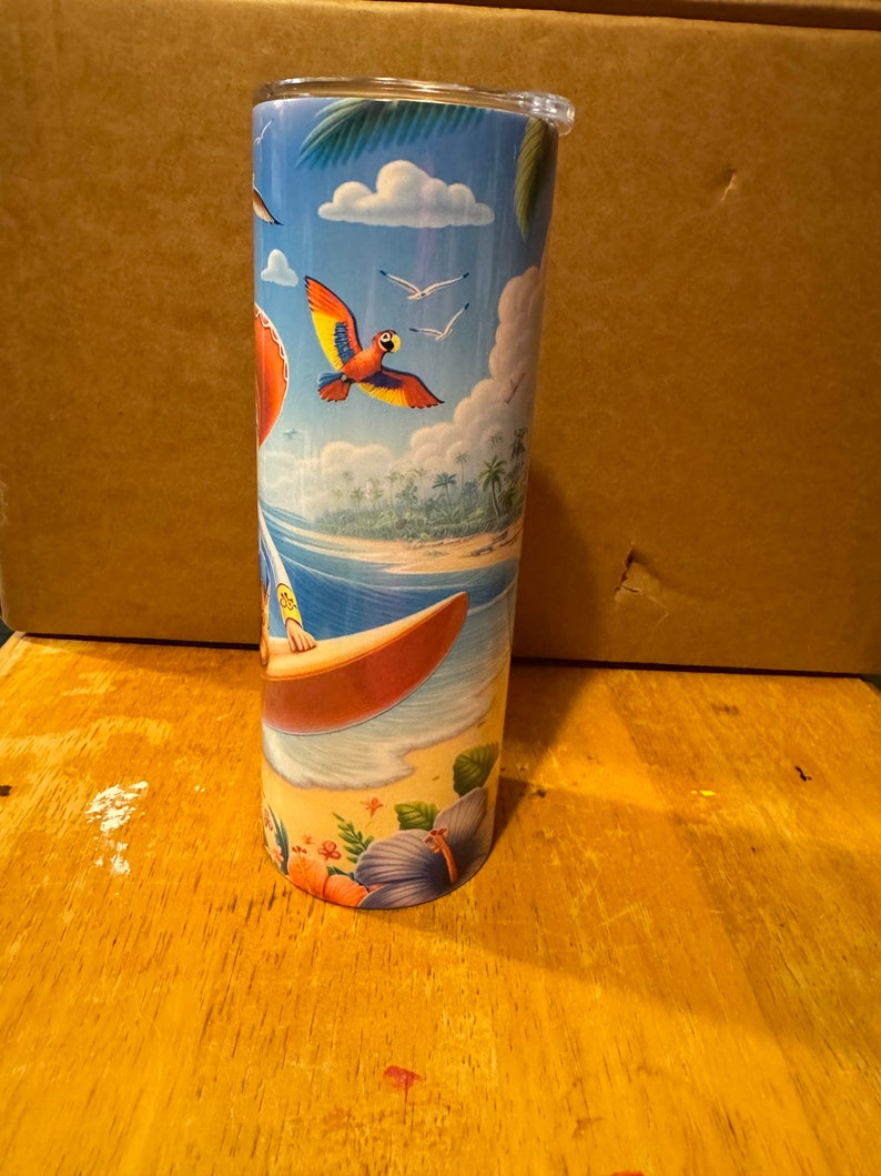 May include: A stainless steel tumbler with a colorful tropical beach scene. The scene features a surfer riding a wave, palm trees, a parrot flying in the sky, and blue water.