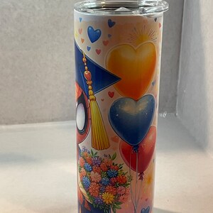 Pode incluir: Um copo de a&ccedil;o inoxid&aacute;vel com um design colorido que apresenta um personagem de desenho animado usando um chap&eacute;u de formatura e segurando um buqu&ecirc; de flores. O design tamb&eacute;m inclui cora&ccedil;&otilde;es, bal&otilde;es e estrelas.