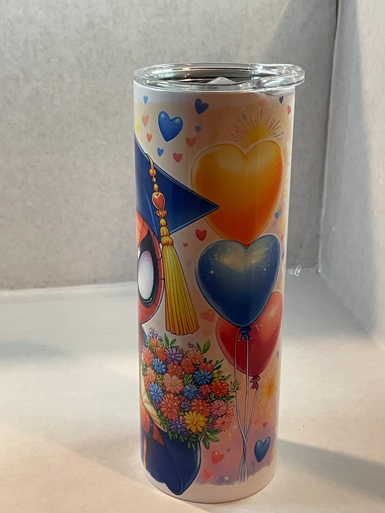 Pode incluir: Um copo de a&ccedil;o inoxid&aacute;vel com um design colorido que apresenta um personagem de desenho animado usando um chap&eacute;u de formatura e segurando um buqu&ecirc; de flores. O design tamb&eacute;m inclui cora&ccedil;&otilde;es, bal&otilde;es e estrelas.