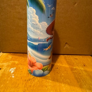May include: A tall, stainless steel tumbler with a colorful design featuring a cartoon character in a sombrero, palm trees, and ocean waves. The tumbler has a clear lid and a pink hibiscus flower design at the bottom.