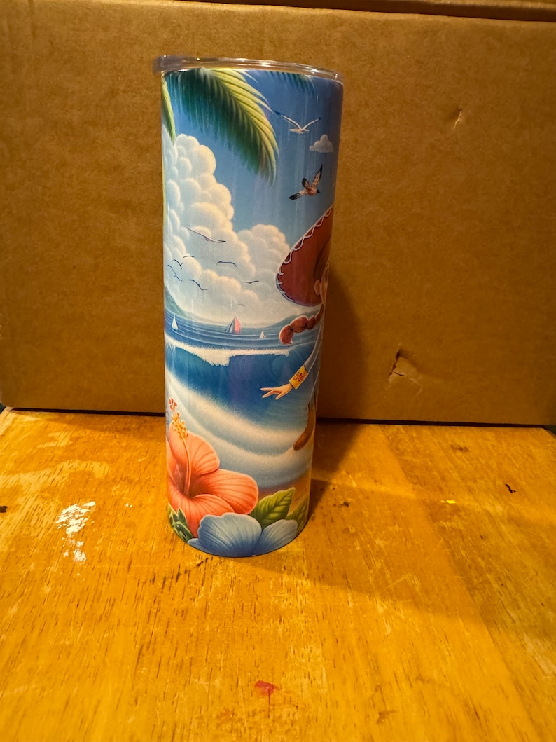 May include: A tall, stainless steel tumbler with a colorful design featuring a cartoon character in a sombrero, palm trees, and ocean waves. The tumbler has a clear lid and a pink hibiscus flower design at the bottom.