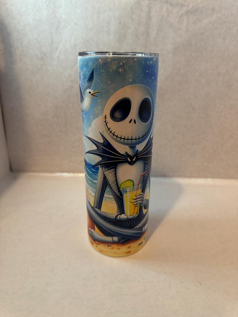 May include: A stainless steel tumbler with a colorful image of Jack Skellington from The Nightmare Before Christmas enjoying a tropical drink on a beach. The background is a blue sky with clouds and a seagull.