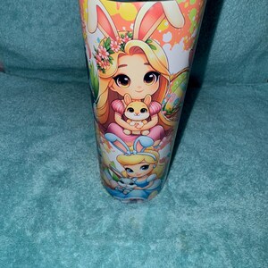 May include: A stainless steel travel mug with an Easter design. The mug features cartoon illustrations of a girl with bunny ears holding a small bunny, and another girl in a blue dress. The background has a speckled pattern.