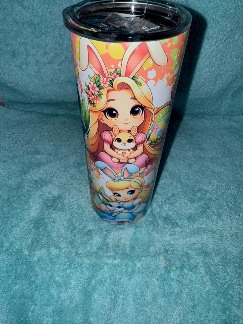 May include: A stainless steel travel mug with an Easter design. The mug features cartoon illustrations of a girl with bunny ears holding a small bunny, and another girl in a blue dress. The background has a speckled pattern.