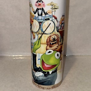 May include: A stainless steel tumbler with a colorful illustration of the Muppets playing music. The Muppets include Kermit the Frog, Animal, Miss Piggy, Fozzie Bear, and a dog. The background is a sheet of music.
