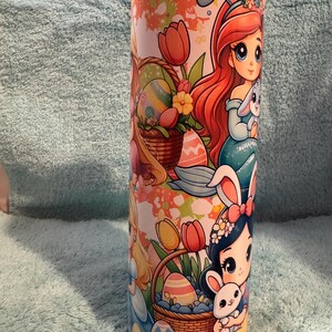 May include: A cylindrical tumbler with a colourful Easter-themed design. The artwork features cartoon princesses, Easter eggs, and bunnies. The tumbler's design incorporates a mix of pastel and bright colours, creating a cheerful aesthetic.