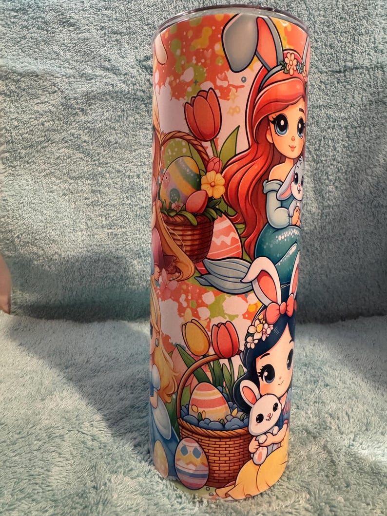 May include: A cylindrical tumbler with a colourful Easter-themed design. The artwork features cartoon princesses, Easter eggs, and bunnies. The tumbler's design incorporates a mix of pastel and bright colours, creating a cheerful aesthetic.