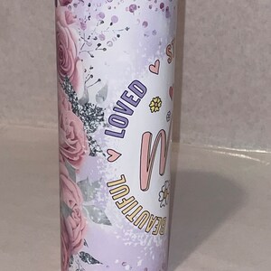 May include: A stainless steel tumbler with a purple and pink floral design. The tumbler has the words "Beautiful" and "Loved" on it, along with a pink heart and a pink letter "M".