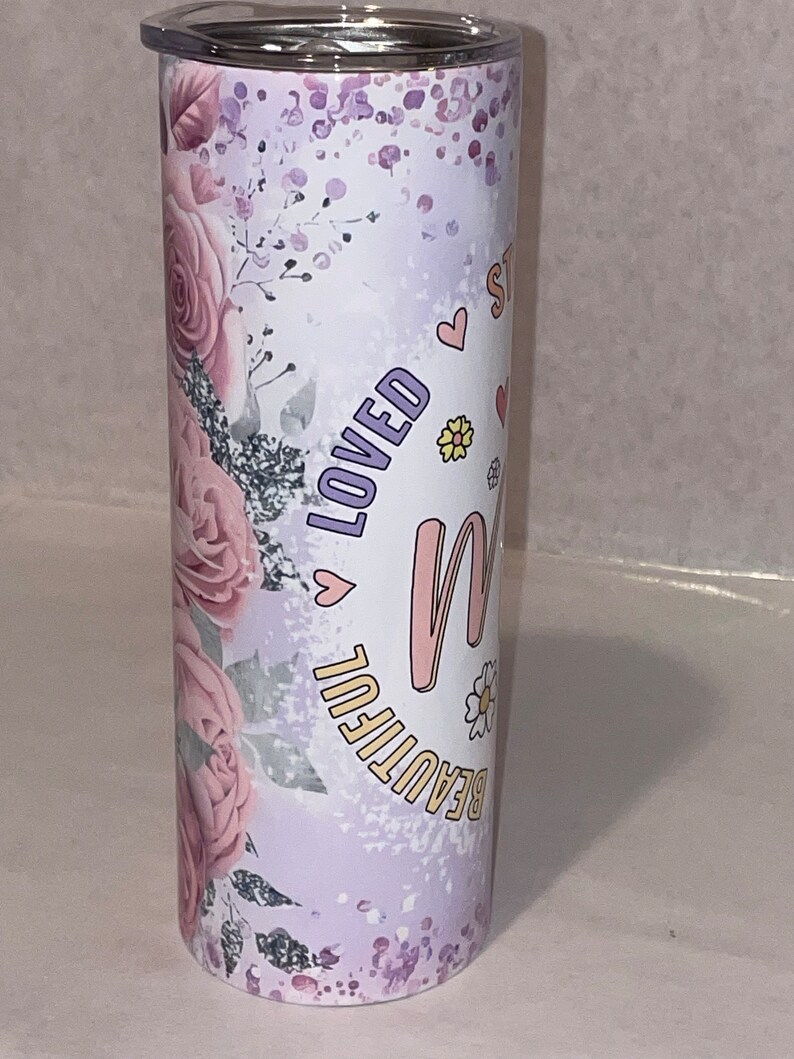 May include: A stainless steel tumbler with a purple and pink floral design. The tumbler has the words "Beautiful" and "Loved" on it, along with a pink heart and a pink letter "M".