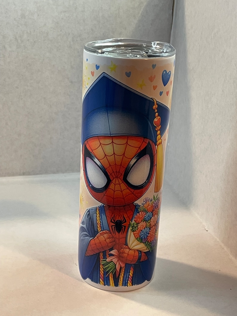 Pode incluir: Um copo de a&ccedil;o inoxid&aacute;vel com um tema de gradua&ccedil;&atilde;o que apresenta uma imagem de desenho animado do Homem-Aranha usando uma touca e uma beca de gradua&ccedil;&atilde;o azuis, segurando um buqu&ecirc; de flores. O fundo &eacute; branco com cora&ccedil;&otilde;es e estrelas azuis, rosa e amarelos.