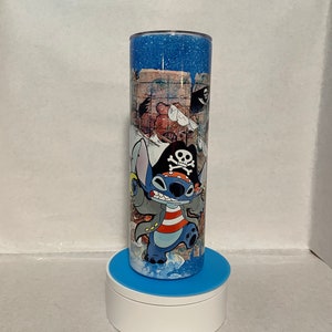 May include: A blue and white stainless steel tumbler with a glitter finish. The tumbler features a cartoon image of Stitch dressed as a pirate. The image includes a treasure map, a skull and crossbones, and a pirate hat.