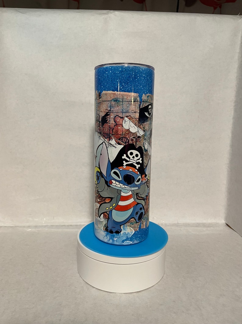 May include: A blue and white stainless steel tumbler with a glitter finish. The tumbler features a cartoon image of Stitch dressed as a pirate. The image includes a treasure map, a skull and crossbones, and a pirate hat.