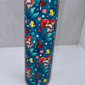 May include: A cylindrical stainless steel tumbler with a teal background featuring a repeating pattern of a red-haired mermaid, a yellow fish, and a red crab, interspersed with white bubbles, pink seashells, and orange starfish, topped with a clear lid.