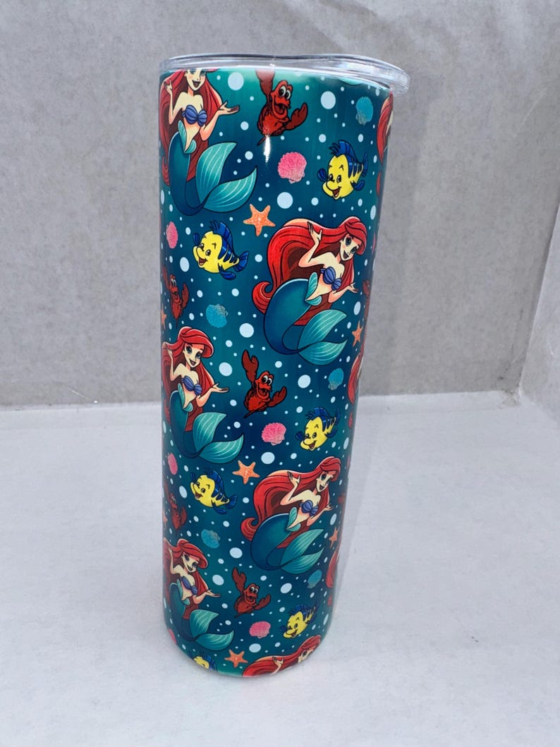 May include: A cylindrical stainless steel tumbler with a teal background featuring a repeating pattern of a red-haired mermaid, a yellow fish, and a red crab, interspersed with white bubbles, pink seashells, and orange starfish, topped with a clear lid.