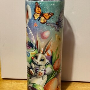 Puede incluir: Un vaso de acero inoxidable con un dise&ntilde;o colorido de Pascua. El dise&ntilde;o presenta un conejito, mariposas, huevos de Pascua y flores. El vaso tiene una tapa negra y mide aproximadamente 20 cm de alto.