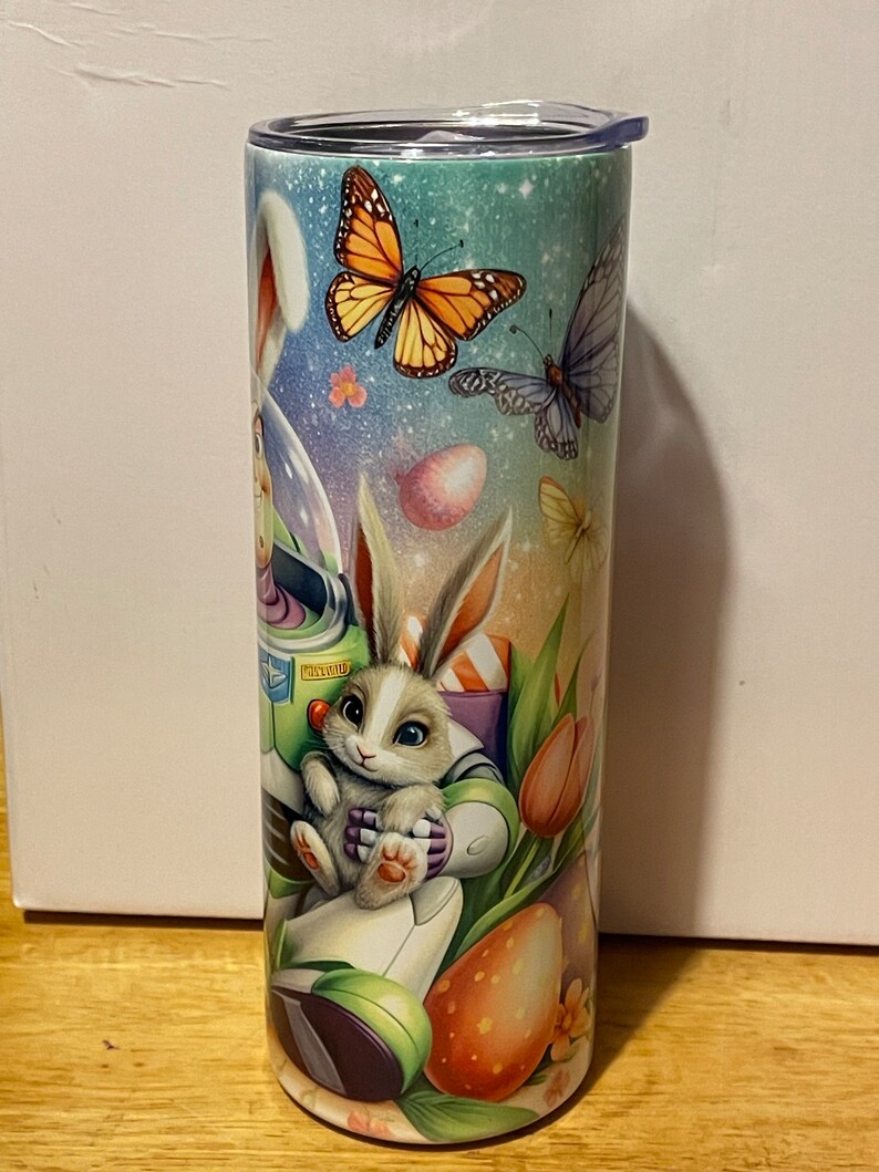 Puede incluir: Un vaso de acero inoxidable con un dise&ntilde;o colorido de Pascua. El dise&ntilde;o presenta un conejito, mariposas, huevos de Pascua y flores. El vaso tiene una tapa negra y mide aproximadamente 20 cm de alto.
