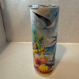 May include: A white stainless steel tumbler with a colorful tropical design. The design features a seagull flying over a beach scene with palm trees, flowers, and a pumpkin.