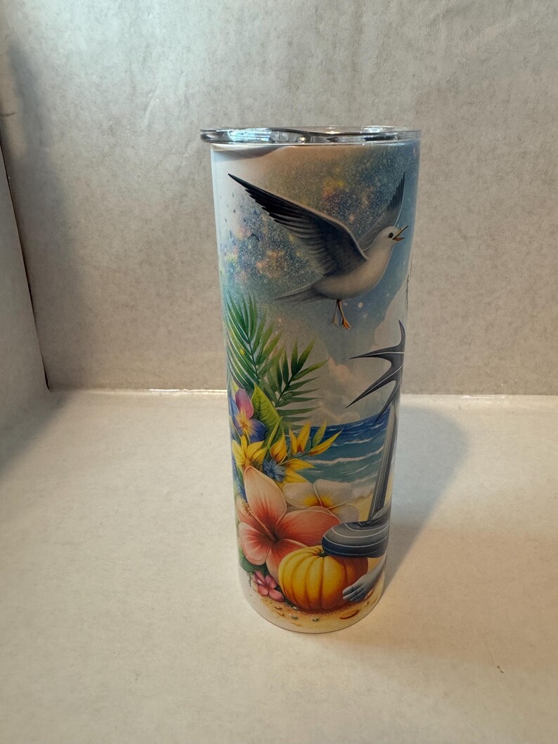 May include: A white stainless steel tumbler with a colorful tropical design. The design features a seagull flying over a beach scene with palm trees, flowers, and a pumpkin.