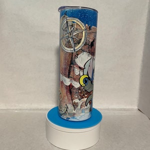 May include: A blue and white tumbler with a nautical theme. The tumbler features a compass, a ship, and a cartoon animal. The tumbler is on a blue and white stand.
