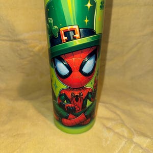 Może przedstawiać: Zielony kubek z postacią z kresk&oacute;wki Spider-Man w zielonym kapeluszu krasnoludka z czarną opaską i klamrą. Kubek ma zielone tło z koniczynami i gwiazdami.