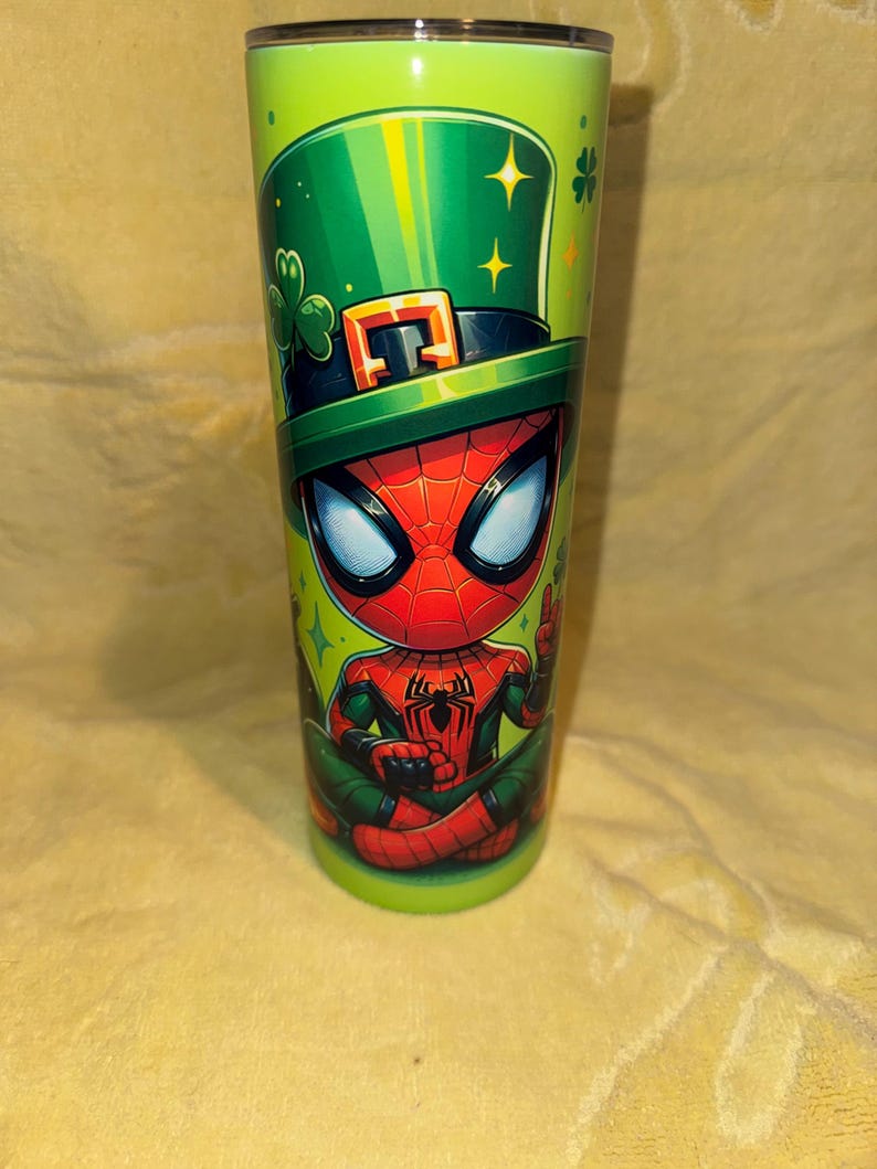 Może przedstawiać: Zielony kubek z postacią z kresk&oacute;wki Spider-Man w zielonym kapeluszu krasnoludka z czarną opaską i klamrą. Kubek ma zielone tło z koniczynami i gwiazdami.