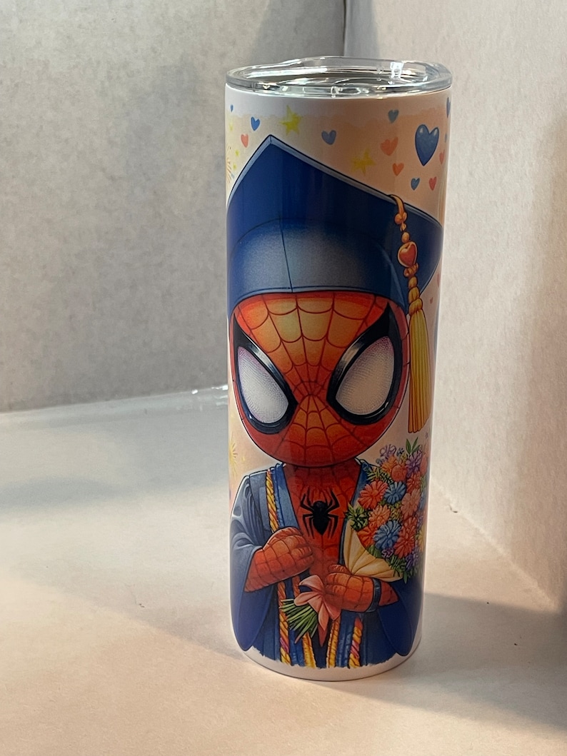 Pode incluir: Um copo de a&ccedil;o inoxid&aacute;vel branco com uma imagem colorida de um Homem-Aranha de desenho animado vestindo um chap&eacute;u de formatura e uma beca. Ele est&aacute; segurando um buqu&ecirc; de flores. O fundo &eacute; amarelo claro com cora&ccedil;&otilde;es e estrelas azuis.