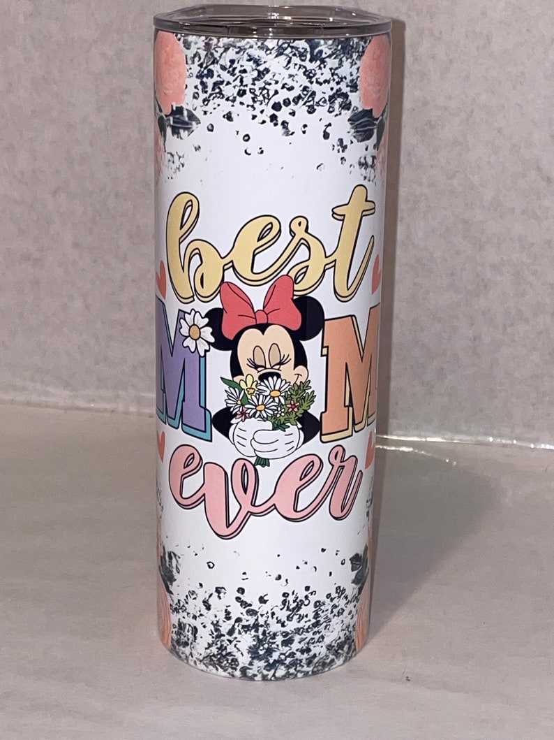 Puede incluir: Un vaso blanco con un fondo moteado en blanco y negro. El vaso presenta una imagen de dibujos animados de Minnie Mouse sosteniendo un ramo de flores. El texto "best mom ever" est&aacute; escrito en letras de colores alrededor de la imagen.