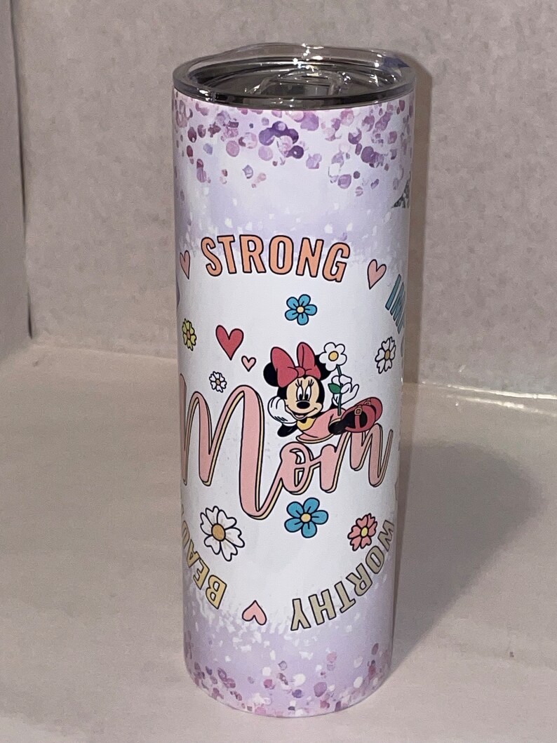 May include: A white stainless steel tumbler with a purple glitter background. It features a pink and white cartoon mouse with a red bow, holding a flower. The text "Strong Mom Worthy Beautiful" is printed around the mouse.