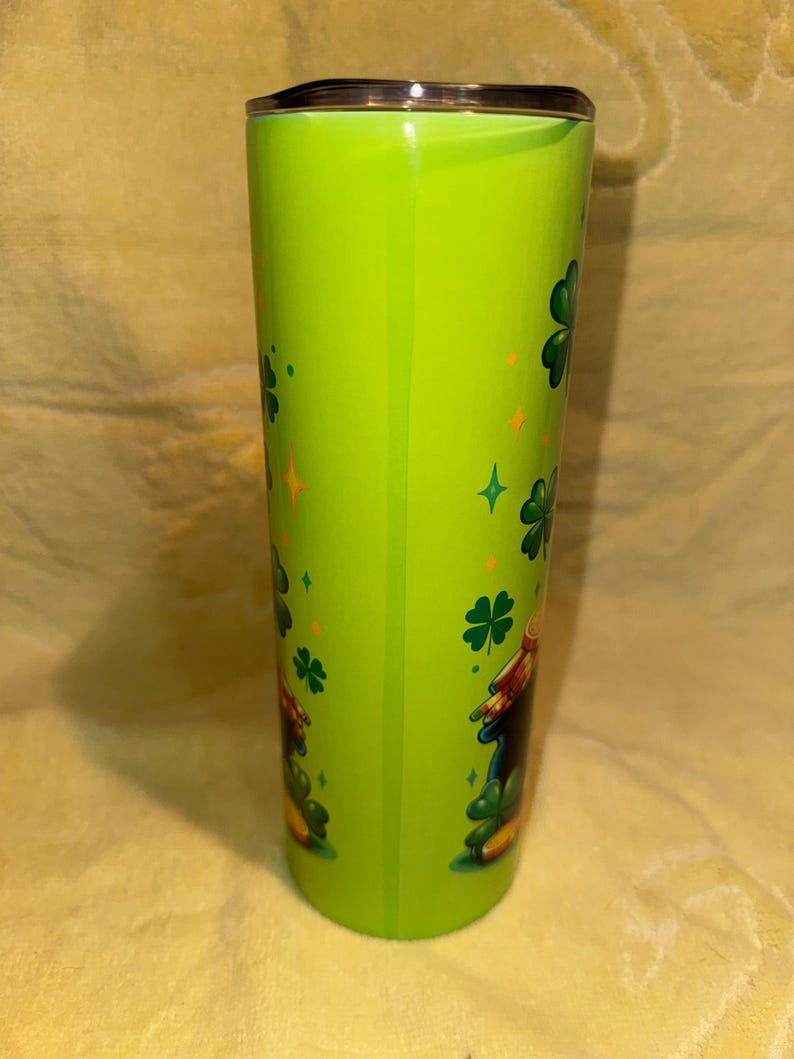 Może przedstawiać: Zielony kubek z czarną pokrywką, ozdobiony koniczyną i złotymi gwiazdami. Kubek ma wz&oacute;r kresk&oacute;wkowego skrzata. Ten izolowany kubek podr&oacute;żny jest idealny do gorących lub zimnych napoj&oacute;w.