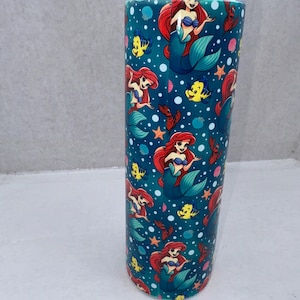 May include: A cylindrical tumbler featuring a repeating pattern of a red-haired mermaid, a yellow fish, and small red crabs on a dark teal background with white bubbles and orange starfish. The drinkware has a clear lid and a smooth, glossy finish.