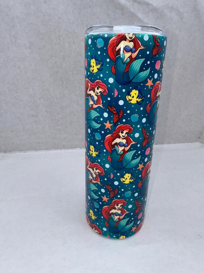 May include: A cylindrical tumbler featuring a repeating pattern of a red-haired mermaid, a yellow fish, and small red crabs on a dark teal background with white bubbles and orange starfish. The drinkware has a clear lid and a smooth, glossy finish.