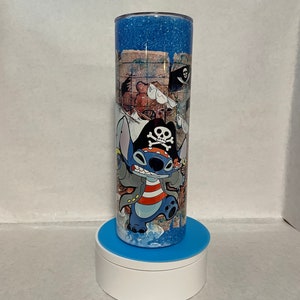 May include: A blue and white tumbler with a glitter finish features a cartoon character wearing a pirate hat and holding a sword. The character is standing on a treasure map with a skull and crossbones flag in the background.