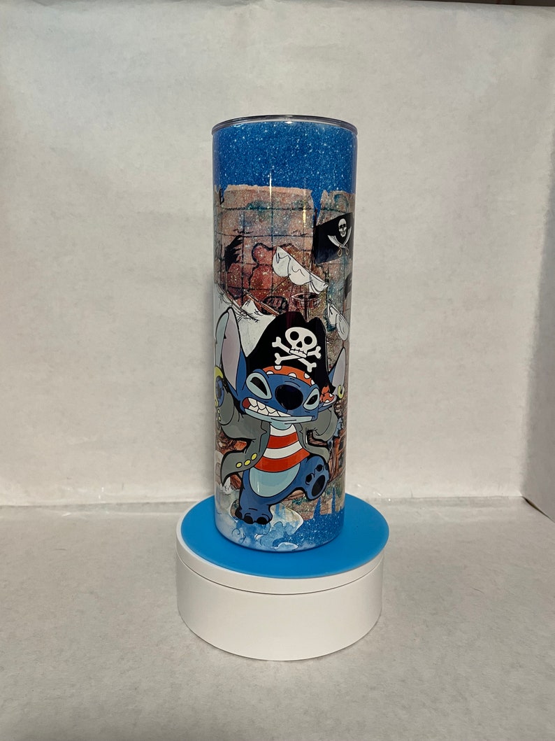 May include: A blue and white tumbler with a glitter finish features a cartoon character wearing a pirate hat and holding a sword. The character is standing on a treasure map with a skull and crossbones flag in the background.