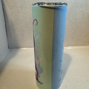 Może przedstawiać: Wysoki, izolowany kubek ze srebrną pokrywką. Kubek podzielony jest na dwie pionowe sekcje: jedną jasnozieloną i drugą jasno niebieską. Zielona sekcja zawiera kolorową ilustrację z r&oacute;żowymi i fioletowymi akcentami.